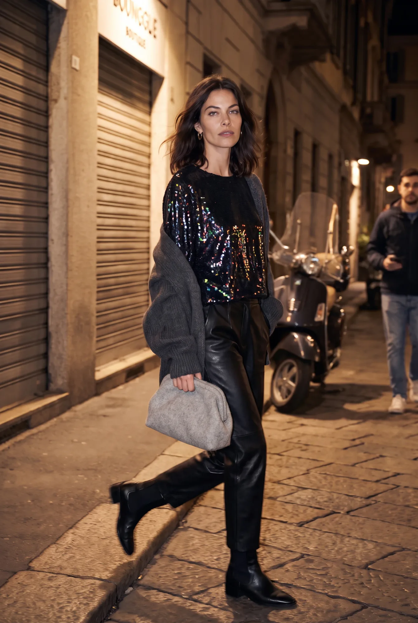 A woman in a black iridescent sequin dolman-sleeve top, grey cardigan, and black leather trousers walks past a scooter on | After the Toast, Relover