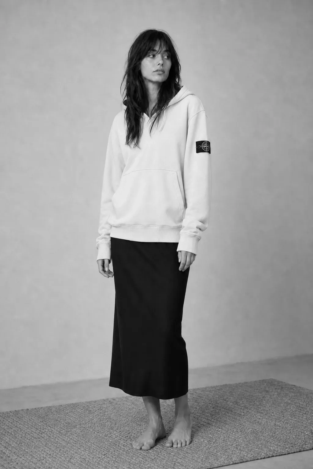 A woman with long dark hair stands in profile-turned pose wearing an ecru pullover hooded sweatshirt with a sleeve patch | The Borrowed Hood, Relover