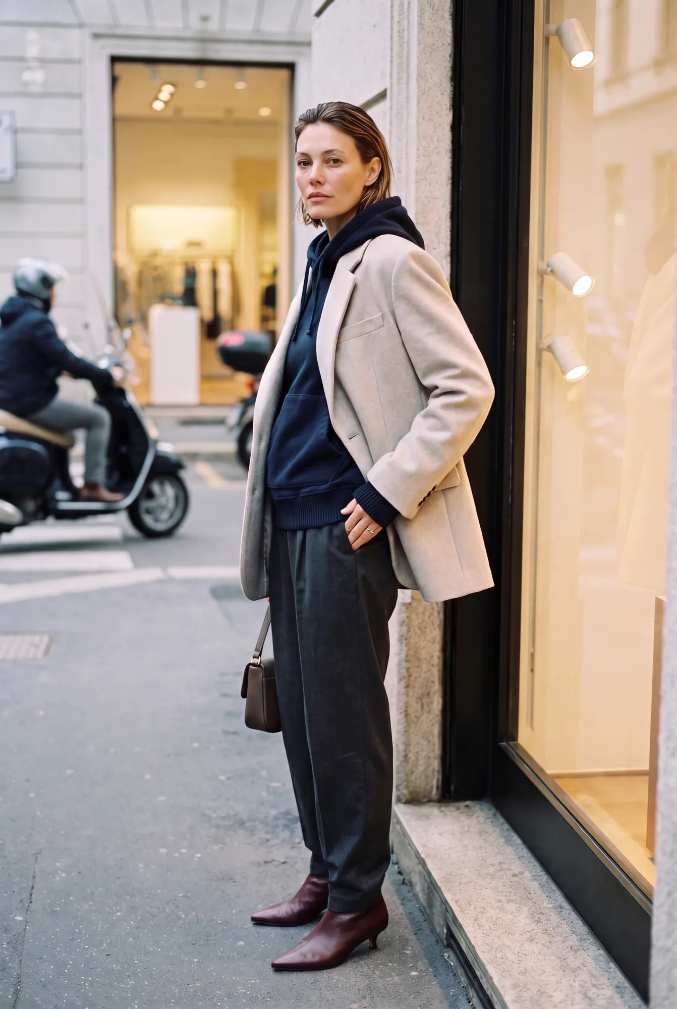 A woman in a beige oversized blazer over a navy pullover hoodie and grey trousers stands outside a shop window beside a | The Borrowed Hood, Relover