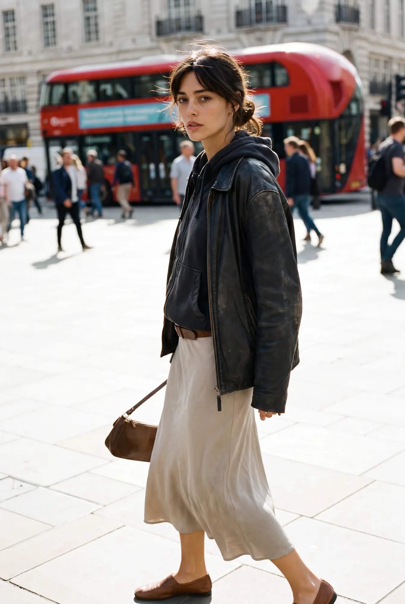 A woman in a worn black leather jacket over a hooded layer and a cream midi skirt walks through a busy plaza with a red | The Borrowed Hood, Relover