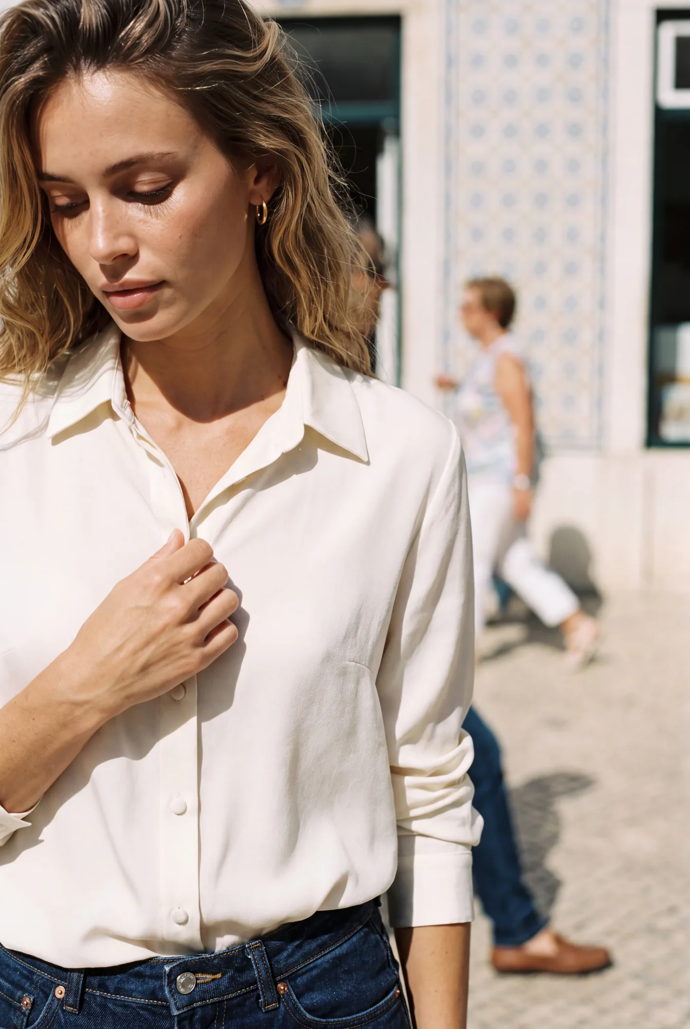 A close-up of a woman wearing an unbuttoned cream silk blouse, touching her collar on a sunlit street | The Dressy Silk Blouse, Relover