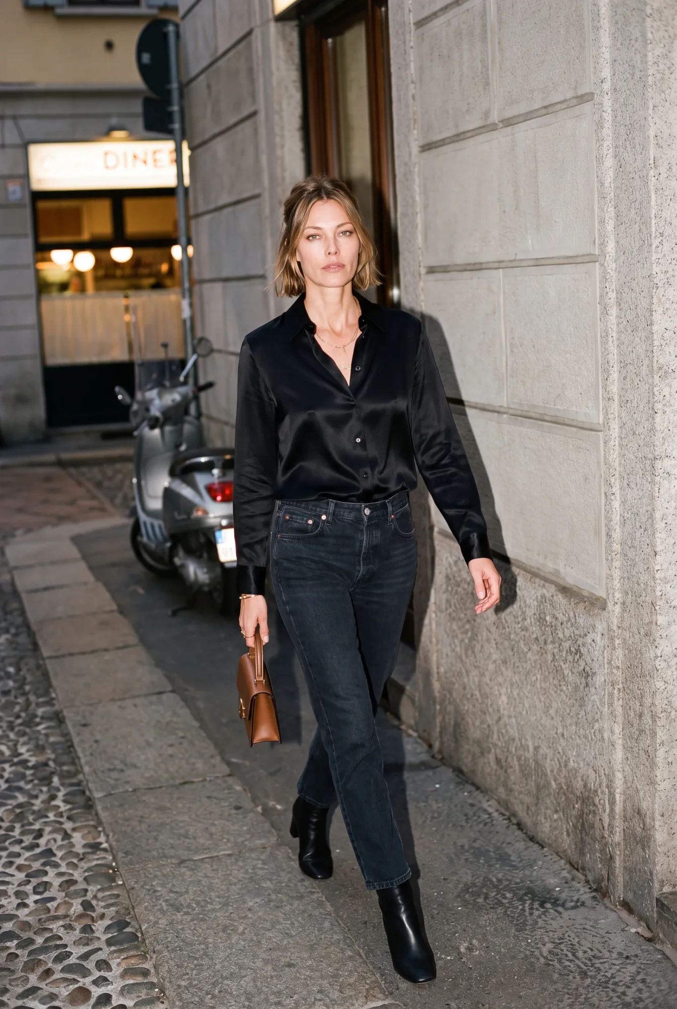 A woman in a black satin blouse and dark jeans walks along a night street past a parked scooter, carrying a small | The Dressy Silk Blouse, Relover