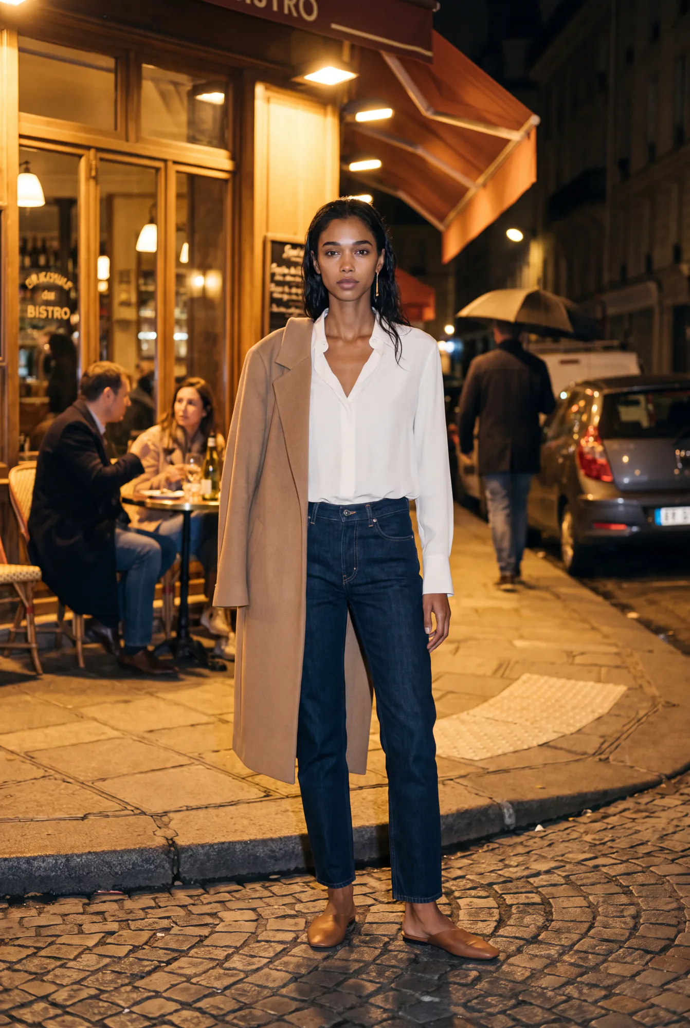 A woman in a white silk blouse and camel coat draped over one shoulder stands on a cobblestone street at night in | The Dressy Silk Blouse, Relover