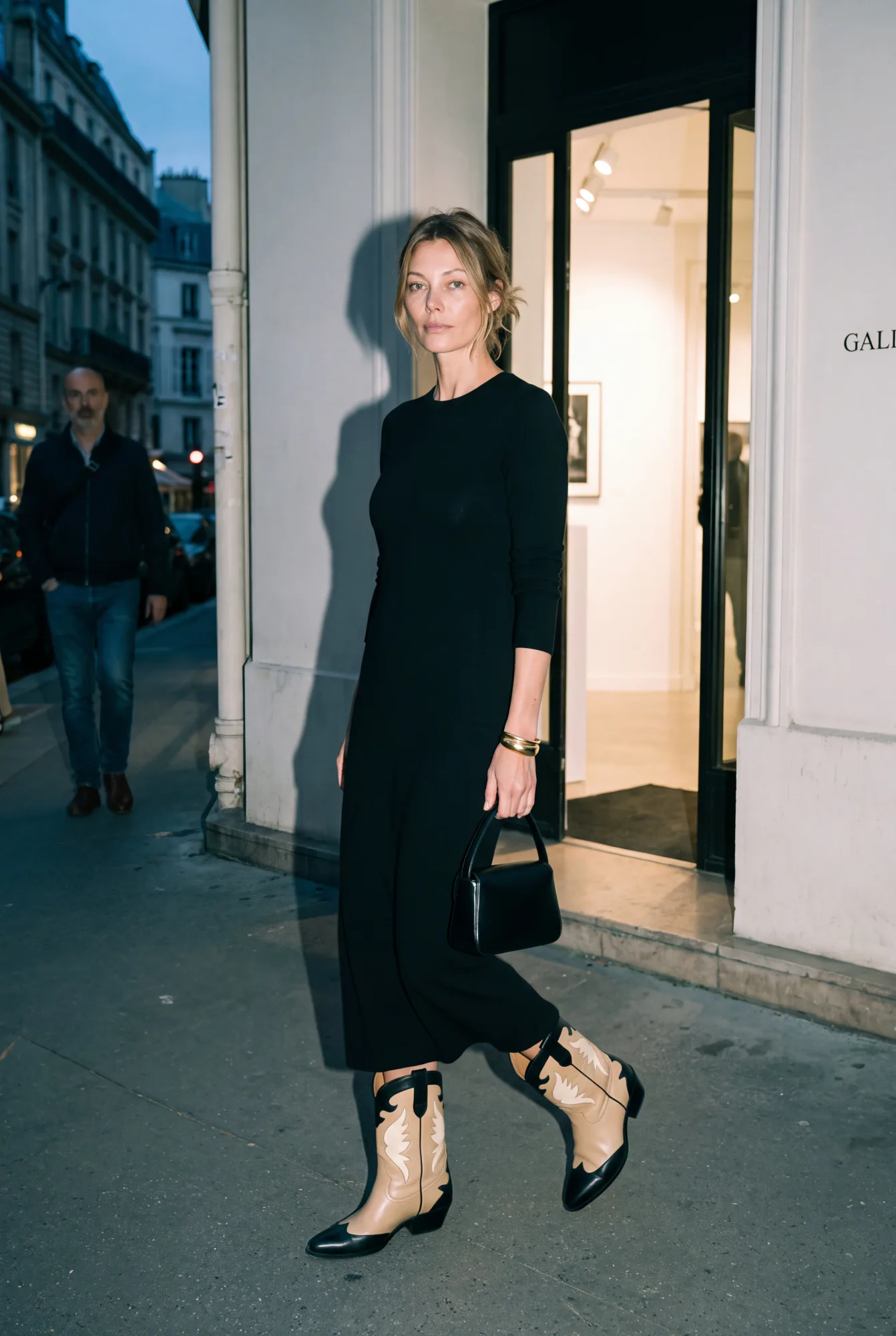 A woman in a black midi dress steps out of a gallery entrance at dusk, wearing beige-and-black two-tone Western boots | The Long Way West, Relover