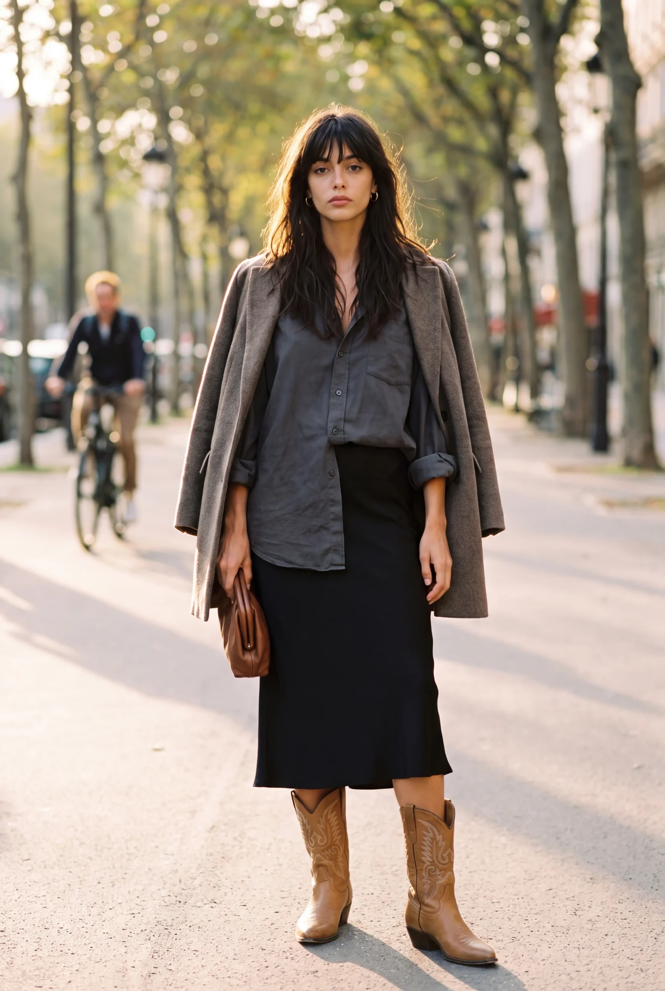 A woman with dark tousled hair and a fringe stands on a tree-lined street in a long coat over a beige silky skirt, wearing tan Western boots | The Long Way West, Relover