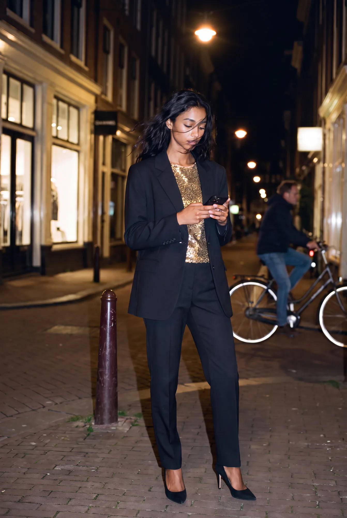 A woman in a long black wool blazer over a gold sequin top and black trousers checks her phone on a cobblestone street | The Quiet Shoulder, Relover
