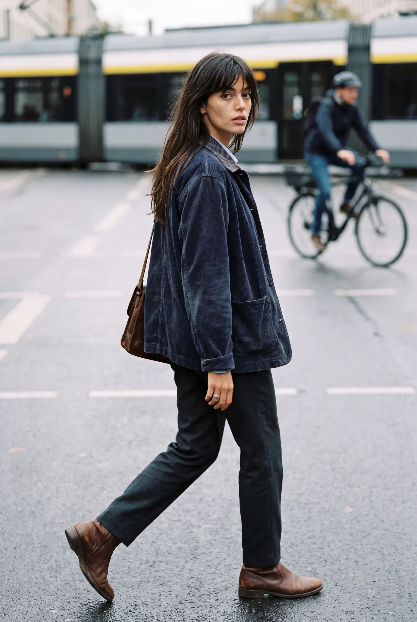 A woman in a navy chore jacket and dark trousers walks across a tram-lined street glancing back over her shoulder, a | The Worker's Layer, Relover