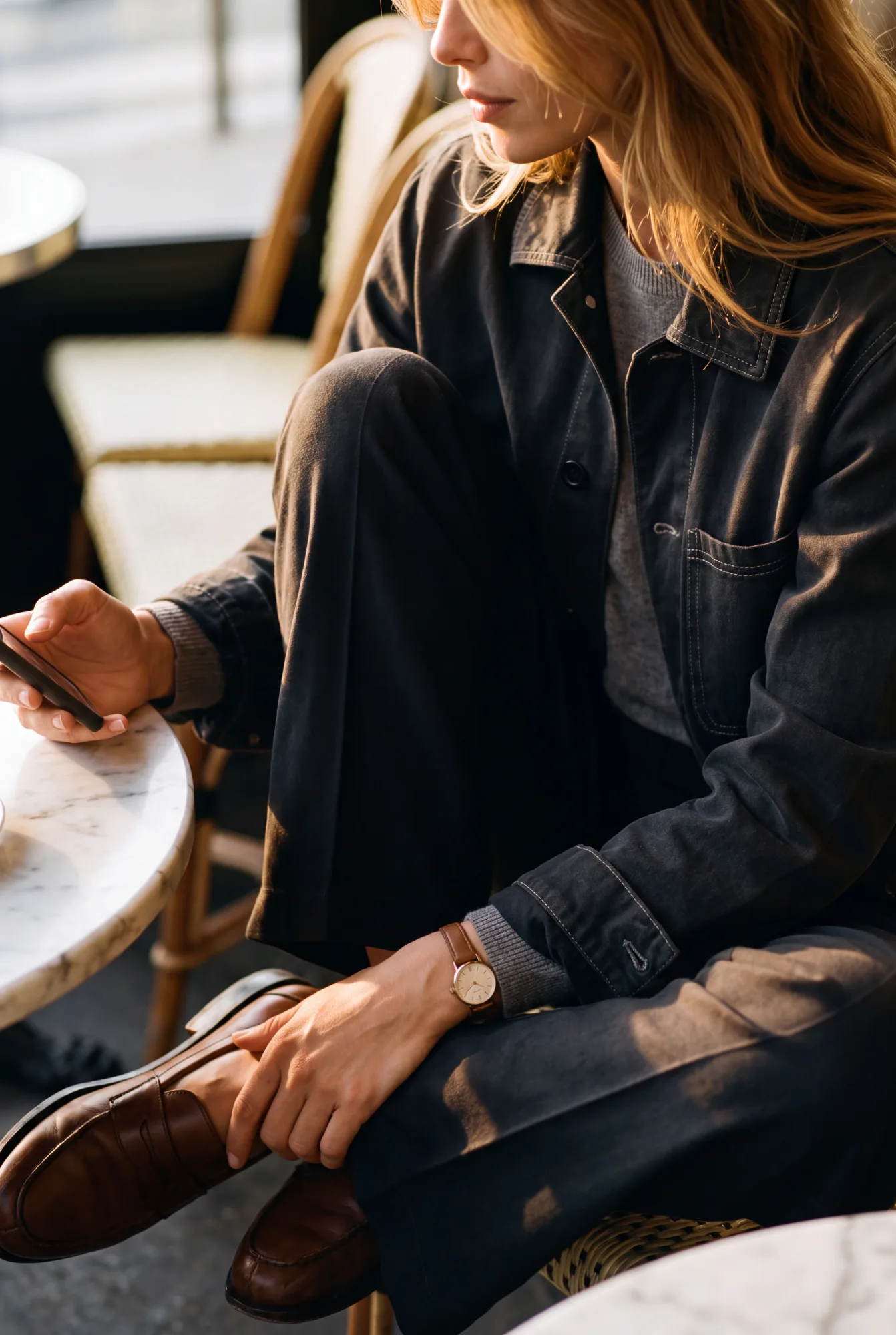A seated woman in a charcoal chore jacket checks her phone at a small cafe table, wearing brown loafers and a wristwatch | The Worker's Layer, Relover