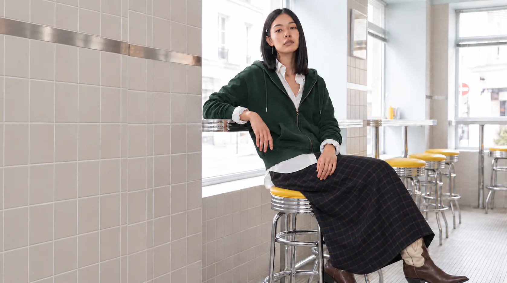 Woman in a dark green zip-up hoodie, checked maxi skirt and two-tone cowboy boots, seated on a yellow café stool.