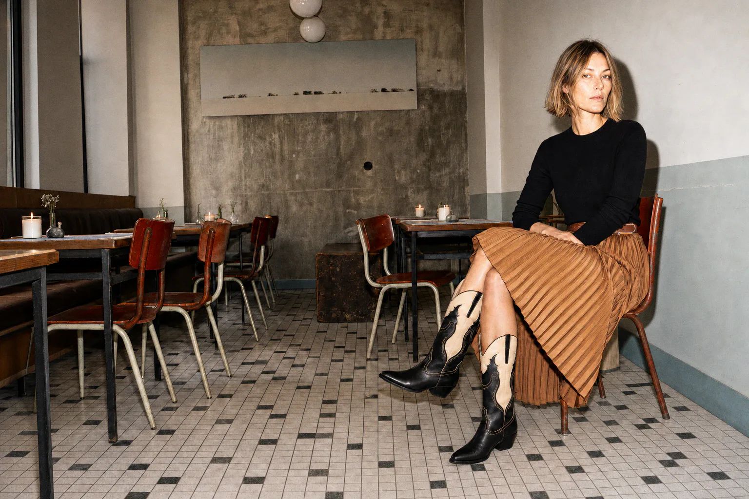 A woman sits cross-legged in a cafe wearing a black top, camel pleated skirt and black-and-beige cowboy boots.